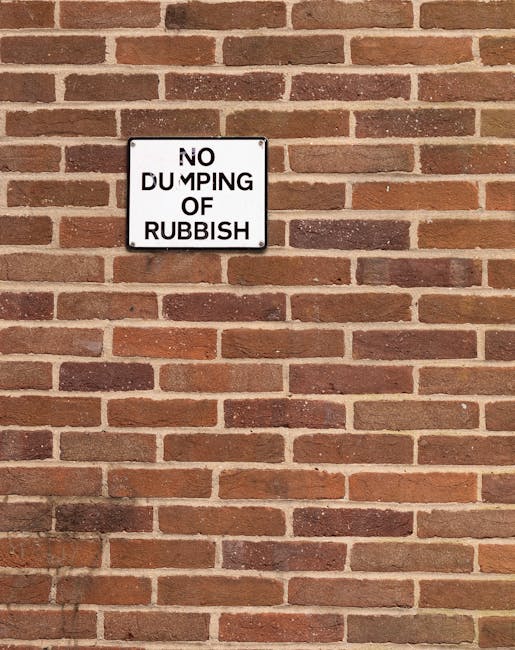 A rectangular white sign with black uppercase letters mounted on a red brick wall. The sign displays the message 'NO DUMPING OF RUBBISH' with some of the text slightly tilted or obscured, possibly due to damage or design. The brick wall features uniformly laid bricks with a reddish-brown hue and light mortar joints in between, creating a textured background. The sign is positioned approximately one-third from the top of the image, and the wall extends evenly across the visible area. The setting suggests an urban or residential boundary where waste disposal regulations are enforced, aligned with private rubbish removal or on-site clearance services. The overall scene has natural outdoor lighting, highlighting the textures of the bricks and the contrast of the sign’s stark white background against the wall, emphasizing the importance of proper waste management to prevent illegal dumping.