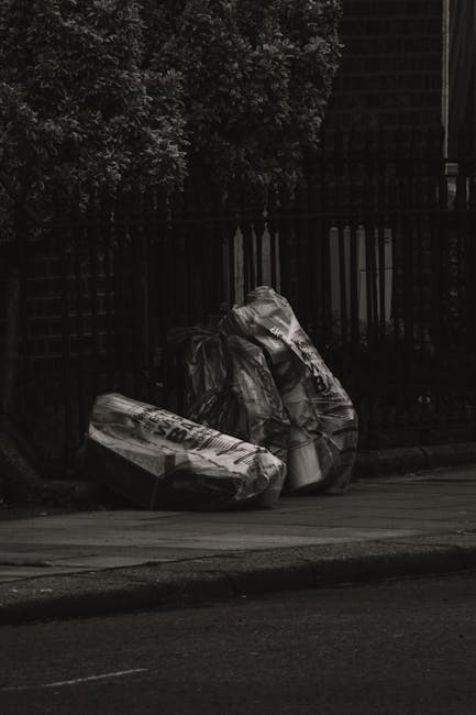 Two large black rubbish bags, made of plastic and tightly knotted at the top, are placed on the curbside on a paved street. The bags appear to contain household waste and have visible text and branding printed on their sides. They are positioned next to a black metal fence with vertical bars, behind which there are dense bushes or small trees with dark foliage. The scene is captured in natural daylight, with a portion of the street surface and a roadside curb visible in the foreground. In the background, there is a wooden or metal structure, possibly a shed or building, partially obscured by the fence and foliage. The scene reflects a typical informal rubbish disposal area, often associated with private waste collection services, with the rubbish bags left for collection by a waste removal company such as Rubbish Removal Kentish Town, in line with alternative waste handling options outside municipal collection.