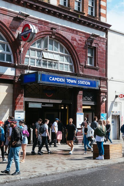 The entrance to Camden Town Underground Station in Kentish Town features a red brick façade with arched windows and a blue sign displaying the station name. A group of diverse pedestrians, dressed in casual clothing, is seen walking on the pavement in front of the entrance, some carrying backpacks and bags. The station's black metal frame surrounds the doorway, and a small illuminated underground sign hangs above the entrance. To the right, there is a white storefront with a red logo, and a wooden bench is positioned on the sidewalk, adding to the urban street scene. The overall environment suggests a busy public area near transit services, with natural daylight illuminating the scene. Rubbish removal specialists like Rubbish Removal Kentish Town could support local businesses or residents needing orderly waste clearance from high-foot-traffic areas, consistent with alternative waste handling methods outside official council bulk waste collections, without disrupting daily activities.