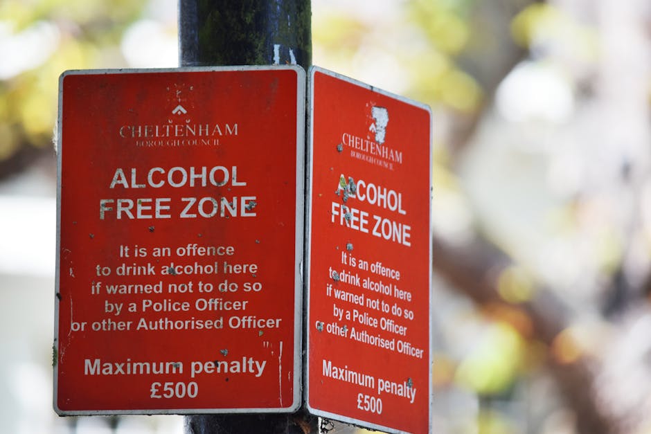 A pair of vertical, rectangular red metal signs attached to a black metal pole in an outdoor setting. The signs indicate a designated alcohol-free zone in Cheltènham, with white text stating that drinking alcohol there is an offence if warned not to do so by a Police Officer or other authorised officer, and that the maximum penalty is £500. The signs show signs of weathering, with some paint chipped and minor rust spots visible. They are positioned on a pavement or street, with a blurred background of leaves and light, suggesting a park or street environment. The signs are part of a censorship for public spaces, often associated with local regulations or community guidelines, and are relevant to the context of managing urban waste and maintaining cleanliness around public areas. Rubbish Removal Kentish Town manages waste around similar signage, providing services for private disposal and on-site clearance.