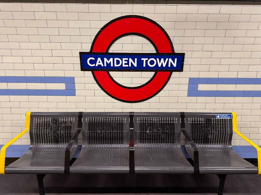 The image depicts a seating area within the Camden Town underground station, featuring four metallic benches with perforated surfaces arranged in a row. The benches are supported by black metal frames, with yellow armrests on either end of the row. Behind the seating, there is a white tiled wall with subtle horizontal blue decorative lines. Above the benches, a prominent circular London Underground sign displays a red outer ring, a blue bar in the middle, and the station name 'Camden Town' written in white text on the blue background. The lighting is neutral, evenly illuminating the scene, and no other objects or environmental elements are visible in the immediate vicinity. The station setting suggests a typical public transport environment, which may be relevant when considering options for private waste disposal or rubbish removal services in the area. Rubbish Removal Kentish Town operates nearby for independent collection and waste management solutions, although no waste or rubbish is visible in this specific image.