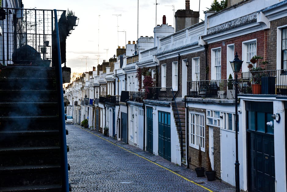 A row of terraced residential houses along a narrow, cobbled street in Kentish Town, featuring brick facades with white and painted detailing. The buildings have small front balconies with black metal railings, some decorated with potted plants and outdoor furniture. On the left, a dark external staircase with metal railings ascends to an upper level, partially obscuring the view. The street is quiet and clean, with the evening light casting a soft glow over the scene. The overall environment suggests a typical urban residential area that may benefit from independent rubbish collection or private waste handling services by companies like Rubbish Removal Kentish Town to manage clutter or clearances efficiently.