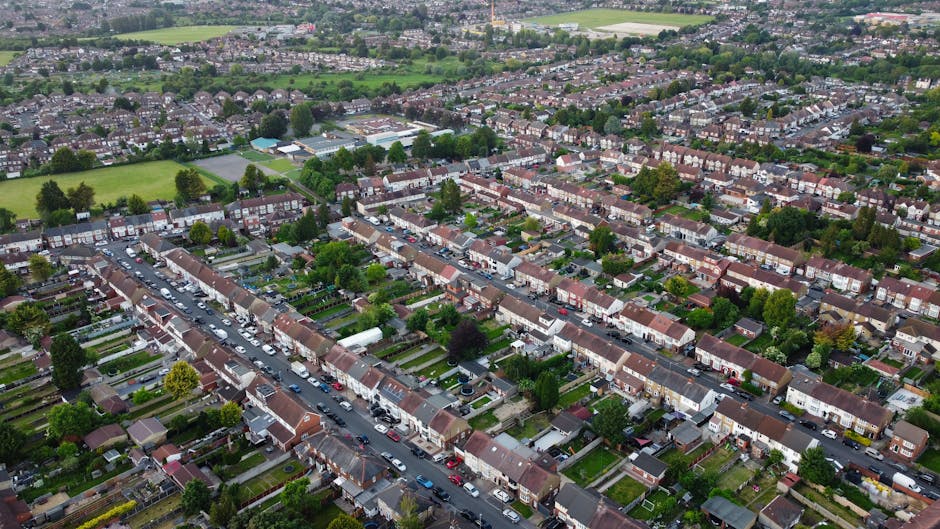 A row of Victorian-era terraced houses situated on a residential street, featuring brick and painted facades, ornate decorative ironwork on the small front balconies, and large bay windows with white frames. The houses are set behind brick retaining walls topped with landscaped gardens and shrubbery. At the street level, there are black wheelie bins lined up along the pavement, indicating waste disposal. The sky above is partly cloudy with patches of blue, and the scene is illuminated by daylight, capturing a quiet neighborhood environment. This setting reflects typical urban housing with potential for local rubbish collection or private waste handling, exemplifying the types of properties that might require rubbish removal services like those offered by Rubbish Removal Kentish Town.