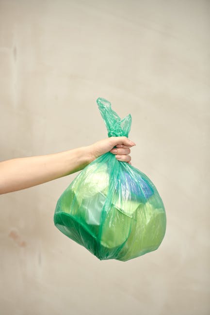 A person’s light-skinned arm is extended into the frame, holding a large green plastic bag filled with waste. The bag appears to contain various discarded materials, possibly packaging or rubbish, and is knotted at the top with a tight twist. The bag's surface is slightly crinkled and shiny, reflecting ambient light. The background is a plain, neutral-colored wall with minimal texture, creating a neutral backdrop that emphasizes the bag and the arm. This image is relevant to rubbish removal services, illustrating the collection of waste in a typical refuse bag, which may be prepared for a private collection or alternative waste handling by companies such as Rubbish Removal Kentish Town, serving clients who need assistance with rubbish clearance from flats or residential properties on Gospel Oak Road NW5 or surrounding areas.