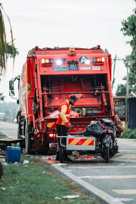 A sanitation worker dressed in a yellow high-visibility vest and dark trousers is standing behind a large red waste collection vehicle on a paved street, actively engaged in waste disposal. The rear compartment of the vehicle is open, revealing a collection chamber with mechanical parts and black hoses, designed for rubbish compaction and transfer. The worker is handling a black plastic bag, which is placed on a small metal platform attached to the vehicle. The scene is set in an urban environment with a sidewalk edged by a patch of grass and trees, and a few commercial signs visible in the background. Overhead, the sky appears partly cloudy with soft natural light illuminating the scene. The waste collection vehicle, likely operated by a private rubbish removal service such as Rubbish Removal Kentish Town, is positioned for on-site waste disposal, highlighting its role in alternative waste handling outside local authority services, especially in urban or densely populated residential settings.