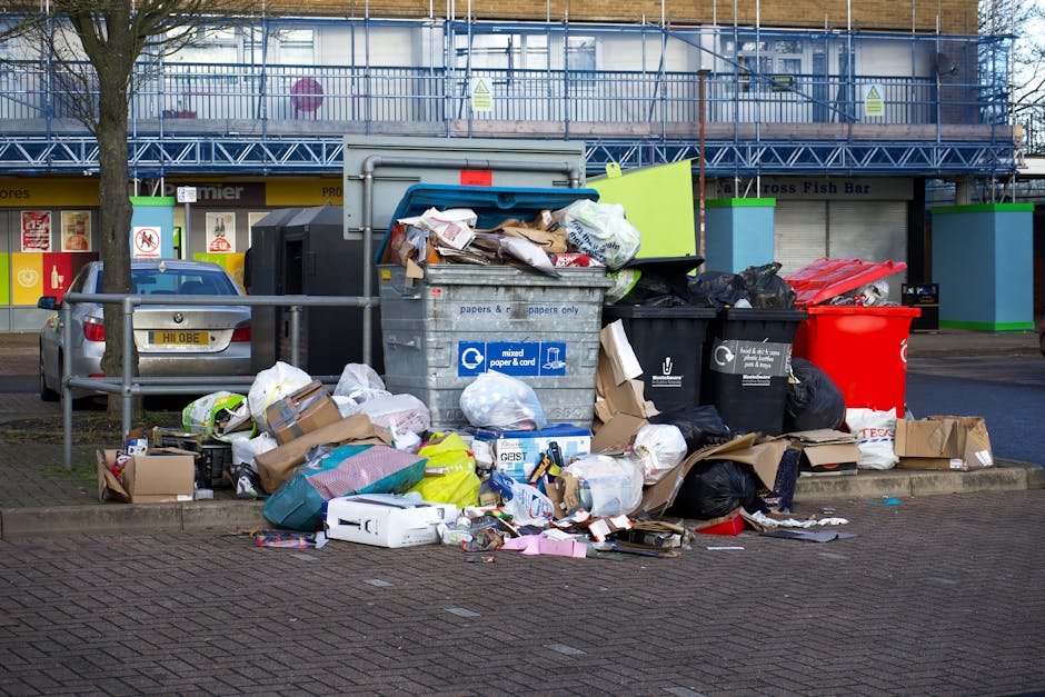 A large collection of mixed household waste, including paper, cardboard, black plastic bags, and miscellaneous packaging, is piled on a paved sidewalk area next to a metal railing. The waste is overflowing from multiple refuse bins, with some rubbish spilling onto the surrounding ground. The bins include a grey bin designated for mixed paper and card, a black bin for general waste, and a bright red bin, which may be for recyclable or hazardous waste. In the background, there is a building with blue scaffolding and protective sheeting, indicating ongoing maintenance or renovation work. To the left, a parked silver car with a UK registration plate is partially visible behind the metal railing, and a tree with a bare trunk is situated nearby. The scene is captured under natural light, highlighting the disorder of the rubbish and contextualizing it within an urban environment that may require private waste removal services such as those provided by Rubbish Removal Kentish Town.