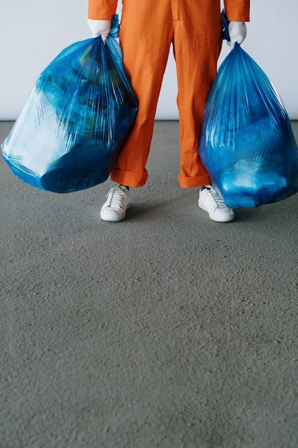 A person standing on a grey carpeted or concrete surface is holding two large, semi-transparent blue plastic rubbish bags, each filled with waste materials. The individual is dressed in orange overalls or work trousers, with only the lower body visible, including white sneakers. The background appears to be an indoor or outdoor environment with a plain, light-colored wall, providing a neutral setting that emphasizes the bags of waste. The bags show amounts of various items indicative of general rubbish, and their plastic material has a slightly crinkled texture with visible folds and creases. The scene reflects a waste collection or rubbish removal activity, likely related to private or independent disposal services such as those offered by Rubbish Removal Kentish Town, aligning with the theme of office waste or general rubbish clearance. The lighting is even, highlighting the sheen of the plastic bags and the subtle texture of the surface beneath, emphasizing the transparent quality of the bags and their contents, suitable for visualising waste collection in a professional context.