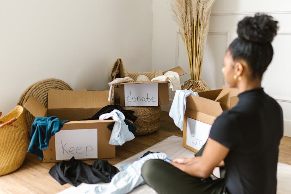 A woman with dark hair styled in a high bun sits on the floor facing away from the camera, in a room with light-colored walls and wooden flooring. In front of her are several open cardboard boxes labeled with handwritten signs reading 'keep,' 'donate,' and 'throw,' containing various types of clothing and textiles. To the left of the boxes, there are woven baskets and a mustard-colored knitted pouf, with some clothing items spilled on the floor. Behind the boxes, tall decorative dried grasses are arranged in a bundle, adding a natural touch to the environment. The scene depicts an organized approach to decluttering or preparing items for disposal, donation, or private disposal services, relevant to professional rubbish removal and recycling options in Kentish Town, as offered by Rubbish Removal Kentish Town, highlighting the sorting process involved in alternative waste handling.