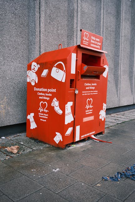 A red donation collection point designed for charitable clothing, book, and household item donations, positioned on a paved sidewalk against a grey concrete wall. The unit features a top slot for depositing donations, a smaller side slot, and a flap opening on the front for collection. The surface is decorated with white icons of clothing, books, handbags, and other personal items, along with text indicating its purpose for donations to the British Heart Foundation. Surrounding the donation point are scattered items and debris, with some discarded fabric or rubbish on the pavement nearby. The scene is outdoors under natural light, emphasizing the functional aspect of the recycling station, which offers an alternative waste handling method for residents seeking to donate unwanted items outside of standard rubbish collection services. This setting aligns with independent or private waste disposal options managed by services such as Rubbish Removal Kentish Town.