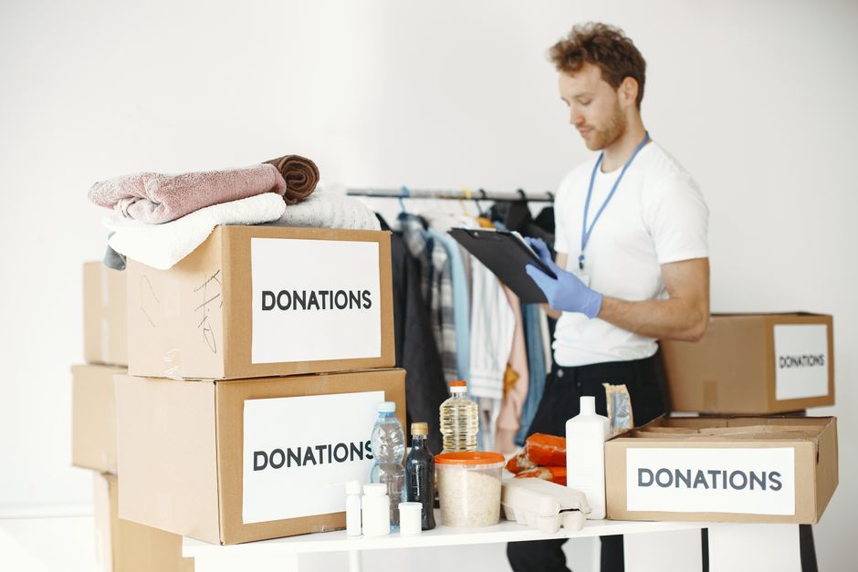 A woman with dark hair styled in a high bun sits on the floor facing away from the camera, in a room with light-colored walls and wooden flooring. In front of her are several open cardboard boxes labeled with handwritten signs reading 'keep,' 'donate,' and 'throw,' containing various types of clothing and textiles. To the left of the boxes, there are woven baskets and a mustard-colored knitted pouf, with some clothing items spilled on the floor. Behind the boxes, tall decorative dried grasses are arranged in a bundle, adding a natural touch to the environment. The scene depicts an organized approach to decluttering or preparing items for disposal, donation, or private disposal services, relevant to professional rubbish removal and recycling options in Kentish Town, as offered by Rubbish Removal Kentish Town, highlighting the sorting process involved in alternative waste handling.