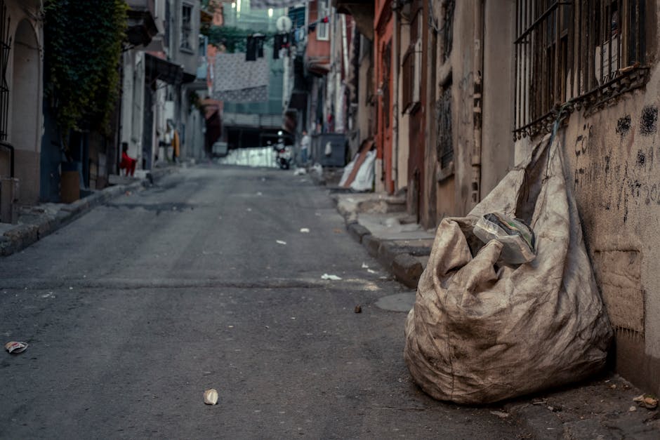 A large, worn, and dirty beige fabric sack filled with assorted rubbish, including a clear plastic bag containing smaller waste items, rests on the pavement against an aged brick wall with black metal security bars on the right side of a narrow, uphill urban alleyway. The street surface appears rough and uneven, with small debris and discarded packaging scattered nearby. On the left side, the alley is lined with tightly packed buildings featuring various doorways, windows, and external fixtures, some with peeling paint and graffiti. In the background, laundry is hung on lines across the street, and a faint outline of buildings and trees can be seen further along the alley. The scene is lit by natural daylight, casting soft shadows, and conveys a typical environment where private waste collection and rubbish removal services like those offered by Rubbish Removal Kentish Town are sometimes required for on-site clearance of waste materials accumulated along urban streets.
