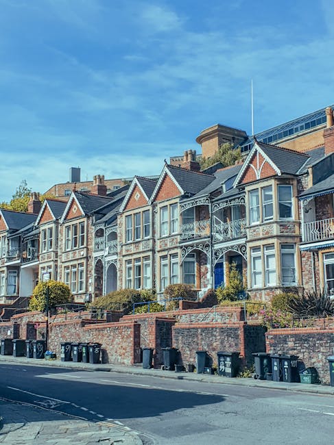 A row of Victorian-style terraced houses with their characteristic bay windows, decorative ironwork, and brick facades in shades of red and brown. The houses are elevated from the street level by brick retaining walls with landscaping including small bushes and plants. The front gardens appear tidy, and the sidewalk runs along a paved road with a few waste bins positioned at the curb. In the background, a modern building with glass windows is visible against a blue sky with some wispy clouds. The scene suggests a residential area where waste collection services, such as those provided by Rubbish Removal Kentish Town, may facilitate alternative waste handling or private rubbish clearance, supporting local living spaces in an urban environment.