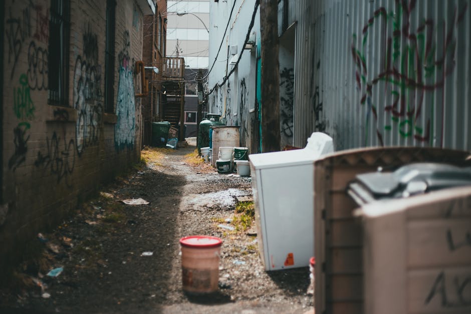 A large, worn, and dirty beige fabric sack filled with assorted rubbish, including a clear plastic bag containing smaller waste items, rests on the pavement against an aged brick wall with black metal security bars on the right side of a narrow, uphill urban alleyway. The street surface appears rough and uneven, with small debris and discarded packaging scattered nearby. On the left side, the alley is lined with tightly packed buildings featuring various doorways, windows, and external fixtures, some with peeling paint and graffiti. In the background, laundry is hung on lines across the street, and a faint outline of buildings and trees can be seen further along the alley. The scene is lit by natural daylight, casting soft shadows, and conveys a typical environment where private waste collection and rubbish removal services like those offered by Rubbish Removal Kentish Town are sometimes required for on-site clearance of waste materials accumulated along urban streets.