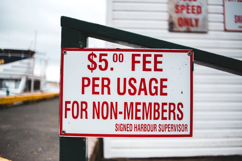 A rectangular white sign with bold red lettering displaying a fee of five pounds per usage for non-members, mounted on a dark green metal post with visible screws at each corner. The sign is situated outdoors near a marina or harbor area, as evidenced by the blurred background showing a portion of boats with white hulls, some with dark accents, docked along a paved surface. To the right of the sign, there is a building with horizontal white siding, and partially visible signs, including one with red text, indicating restrictions or guidelines related to harbor or storage regulations. The lighting appears natural and overcast, casting a soft, even tone over the scene, emphasizing the sign's clarity and the surrounding environment, which suggests a maritime or waste management context associated with private or independent disposal services often necessary for non-members in a harbour setting.
