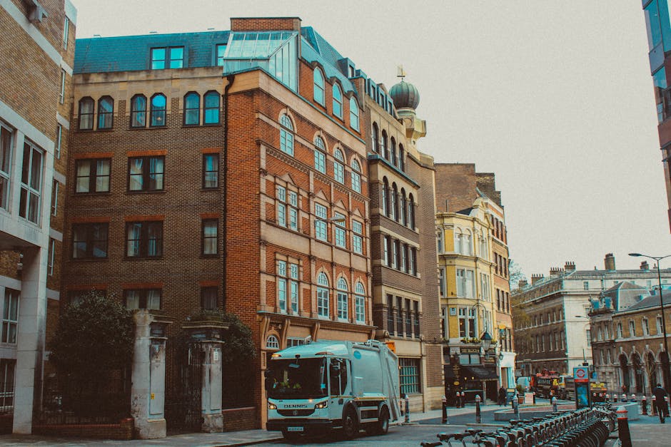 A street scene in an urban area showing multi-story historic buildings with a mix of brick facades and ornate architectural details, including arched windows and decorative cornices. The buildings vary in color, with one prominent structure featuring a red brick exterior, large arched windows, and a blue roof topped with a small decorative dome. In the foreground, a light blue waste collection truck is parked on the paved street, positioned near a gated entrance to a property, with the company's branding visible on the vehicle. To the right, a row of bicycles is secured to a bike rack, and several bollards line the sidewalk, separating pedestrian space from the roadway. The environment appears well-lit by natural daylight, emphasizing the textures of the brickwork and the metallic surface of the waste truck. This scene illustrates the integration of private waste collection services like Rubbish Removal Kentish Town in urban settings, with the waste vehicle ready for roadside rubbish collection or on-site clearance activities in a busy city area.