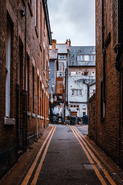 A narrow alleyway bordered by tall brick buildings on both sides, featuring a paved road with double yellow lines running along the edges. In the background, there is a small courtyard area with various discarded items including large black plastic bins, a wooden staircase attached to a multi-storey building, and a graffiti-covered wall. The alley is overcast with cloudy skies, and the environment appears to be an urban setting. This scene illustrates an area where private waste collection services, such as those provided by Rubbish Removal Kentish Town, might operate for clearing accumulated refuse in city backstreets, highlighting the need for efficient rubbish removal in built-up residential or commercial zones. The surface of the alley shows signs of recent use, with some wet patches on the pavement, and the scene overall emphasizes the typical environment for independent rubbish collection or on-site clearance activities in densely populated areas.
