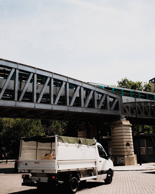The image depicts a white commercial van parked beneath an elevated railway or metro bridge in an urban environment. The van, positioned in the foreground, has an open cargo area covered by a green tarpaulin, with some visible cardboard boxes and debris inside, suggesting recent loading or unloading activity. The vehicle's surface appears to be slightly weathered, typical of work-related vehicles used for waste or rubbish collection. The surrounding area includes a paved surface, likely a sidewalk or small plaza, with a few trees and parts of nearby modern buildings visible in the background. The bridge above is constructed from metal trusses and concrete supports, painted in shades of grey, and spans across the scene with a green train passing overhead, creating a sense of movement. The lighting is bright and natural, indicating daylight conditions. This scene subtly relates to independent waste collection or on-site clearance activities, aligning with services offered by rubbish removal companies such as Rubbish Removal Kentish Town, which handle private waste disposal tasks in busy city environments.