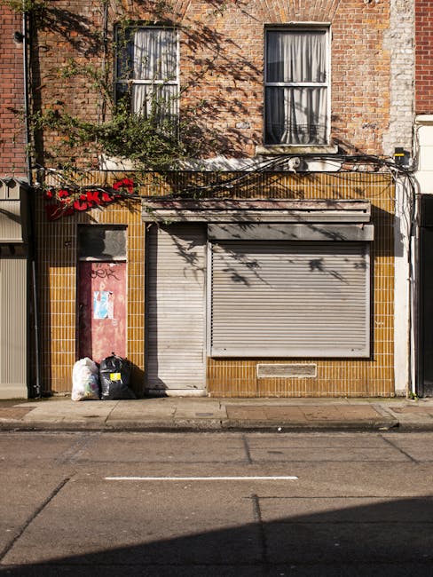 A narrow alleyway bordered by tall brick buildings on both sides, featuring a paved road with double yellow lines running along the edges. In the background, there is a small courtyard area with various discarded items including large black plastic bins, a wooden staircase attached to a multi-storey building, and a graffiti-covered wall. The alley is overcast with cloudy skies, and the environment appears to be an urban setting. This scene illustrates an area where private waste collection services, such as those provided by Rubbish Removal Kentish Town, might operate for clearing accumulated refuse in city backstreets, highlighting the need for efficient rubbish removal in built-up residential or commercial zones. The surface of the alley shows signs of recent use, with some wet patches on the pavement, and the scene overall emphasizes the typical environment for independent rubbish collection or on-site clearance activities in densely populated areas.