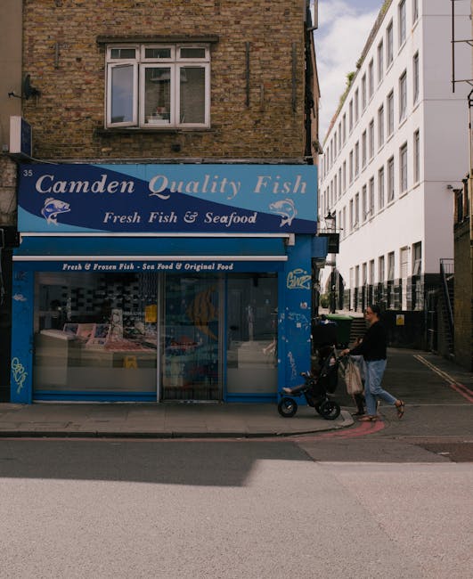The image depicts the exterior of a small fish and seafood shop in Camden, with a blue sign reading 'Camden Quality Fish' and additional signage advertising fresh fish and seafood, as well as frozen options. The shop's front includes a glass display case filled with fish and seafood products, partially visible behind a metal security gate. The building above the shop has a brick facade with a white-framed window on the upper floor. To the right of the shop, in the background, there is a modern white multi-story building with large windows, contrasting with the older brick structure. In the foreground, a person dressed in a black top and light trousers is pushing a stroller along the sidewalk, partially situated on a curb with a red-painted edge, possibly indicating a designated loading or no-parking zone. The street appears quiet with minimal traffic, and natural daylight illuminates the scene, casting soft shadows. This setting showcases a typical urban retail environment with elements that could relate to waste disposal and rubbish removal, especially considering the proximity to diverse building types and potential waste generation areas. Rubbish Removal Kentish Town often services such locations for independent waste collection or on-site clearance efforts.